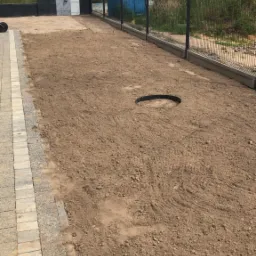 Przygotowany teren pod trawnik z rolki, widoczny okrągły wykop, obok ułożona kostka brukowa, w tle ogrodzenie panelowe i przenośna toaleta.