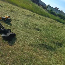 Świeżo skoszona trawa na zboczu z widoczną kosiarką i wysoką trawą w tle, domy w oddali.