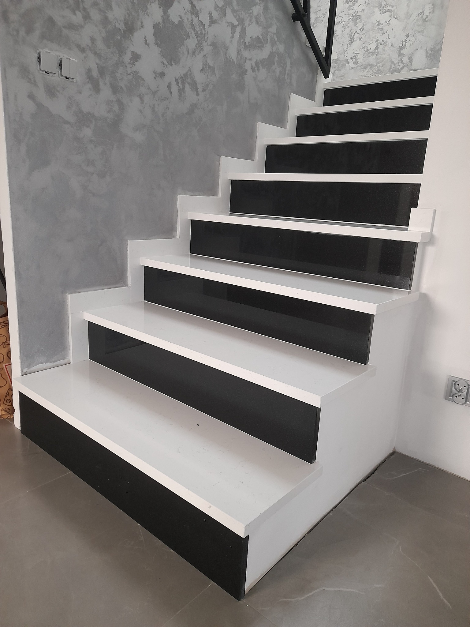 Nowoczesne schody wewnętrzne z białymi stopnicami i czarnymi podstopnicami, ściana z szarą dekoracyjną fakturą, widoczne gniazdko elektryczne.