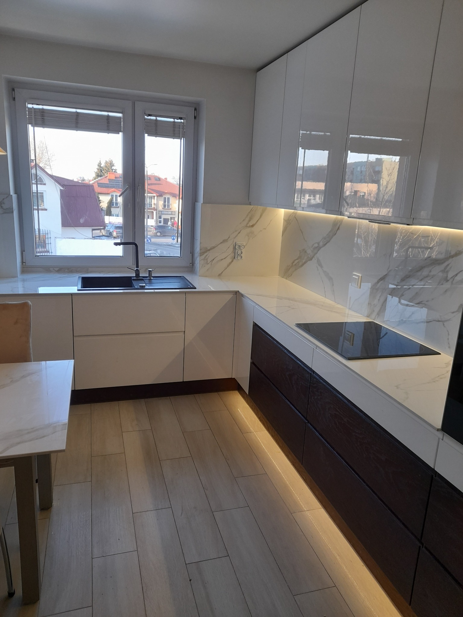Nowoczesna kuchnia z białymi, lakierowanymi szafkami górnymi i ciemnobrązowymi szafkami dolnymi, marmurowy blat i backsplash, czarny zlew pod oknem, oświetlenie LED pod szafkami dolnymi, widok...