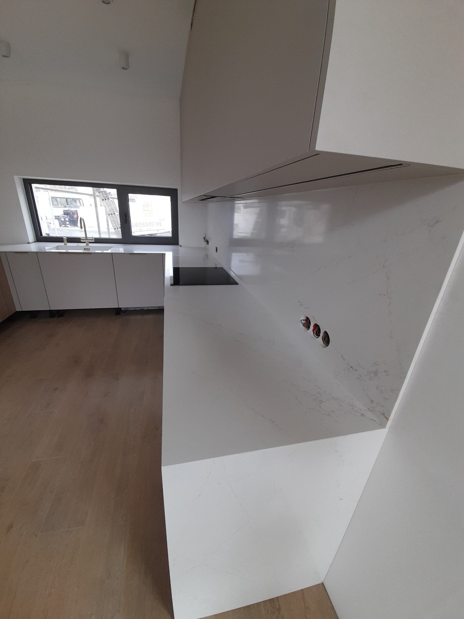 Nowoczesna, minimalistyczna kuchnia z białymi szafkami i blatem z imitacji kamienia, widoczne gniazdka elektryczne w ścianie i płyta indukcyjna w blacie.