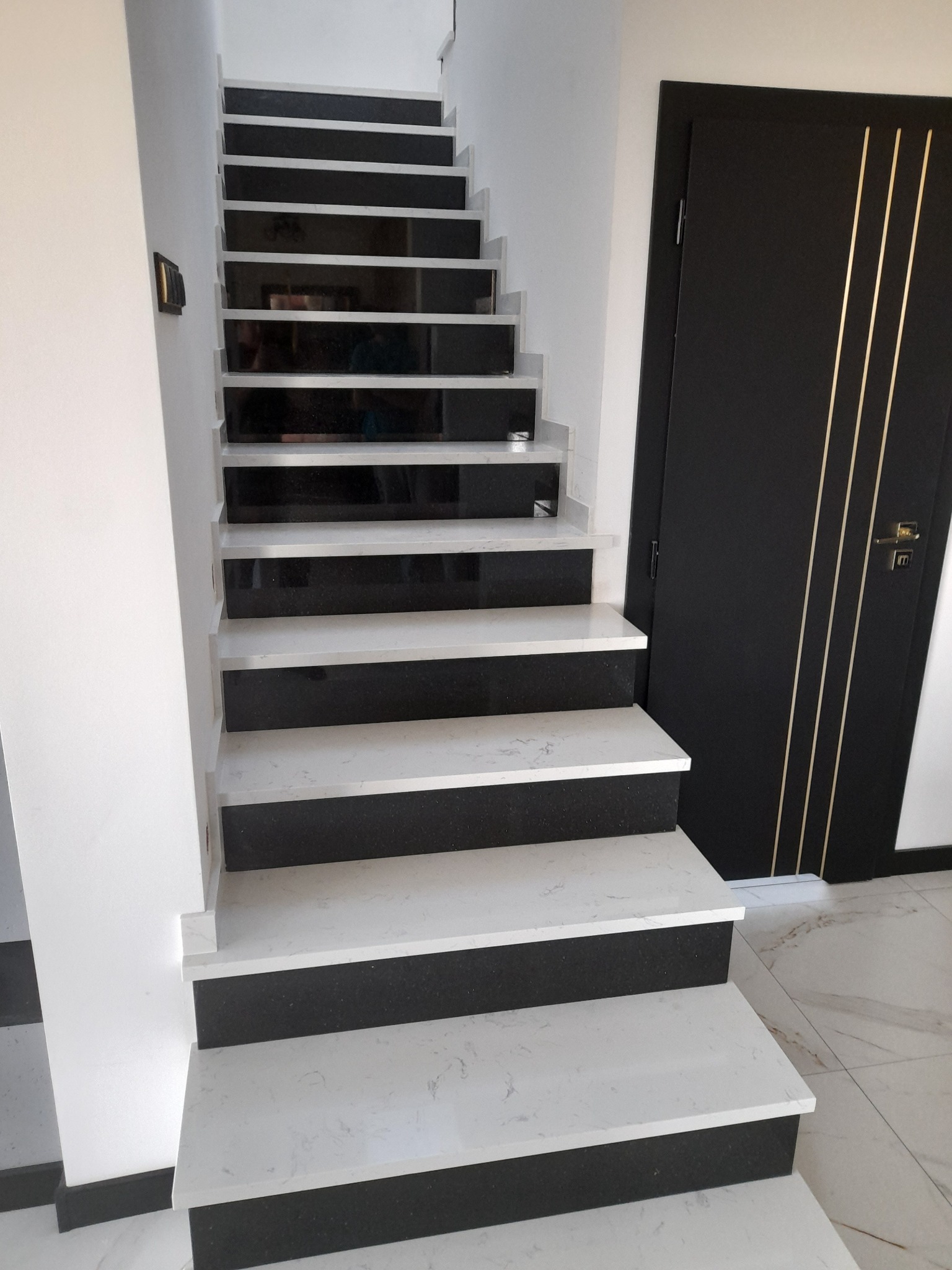 Nowoczesne schody wewnętrzne z białymi stopniami z wzorem marmuru i czarnymi podstopnicami, obok czarne drzwi z złotymi paskami.