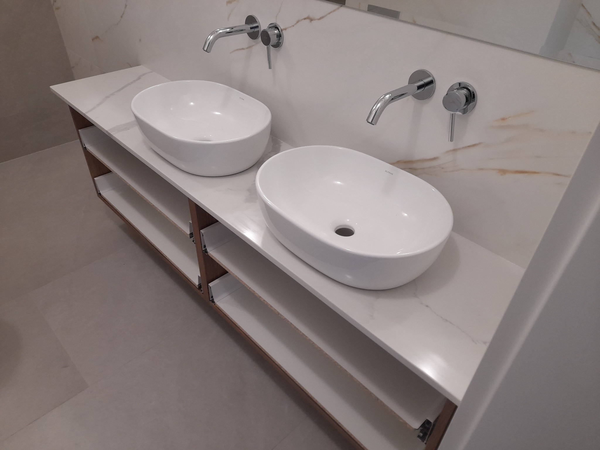 Dwie owalne umywalki nablatowe marki Oltris z chromowanymi bateriami ściennymi, zamontowane na blacie z białego marmuru z otwartymi półkami w łazience wykończonej w jasnych kolorach.