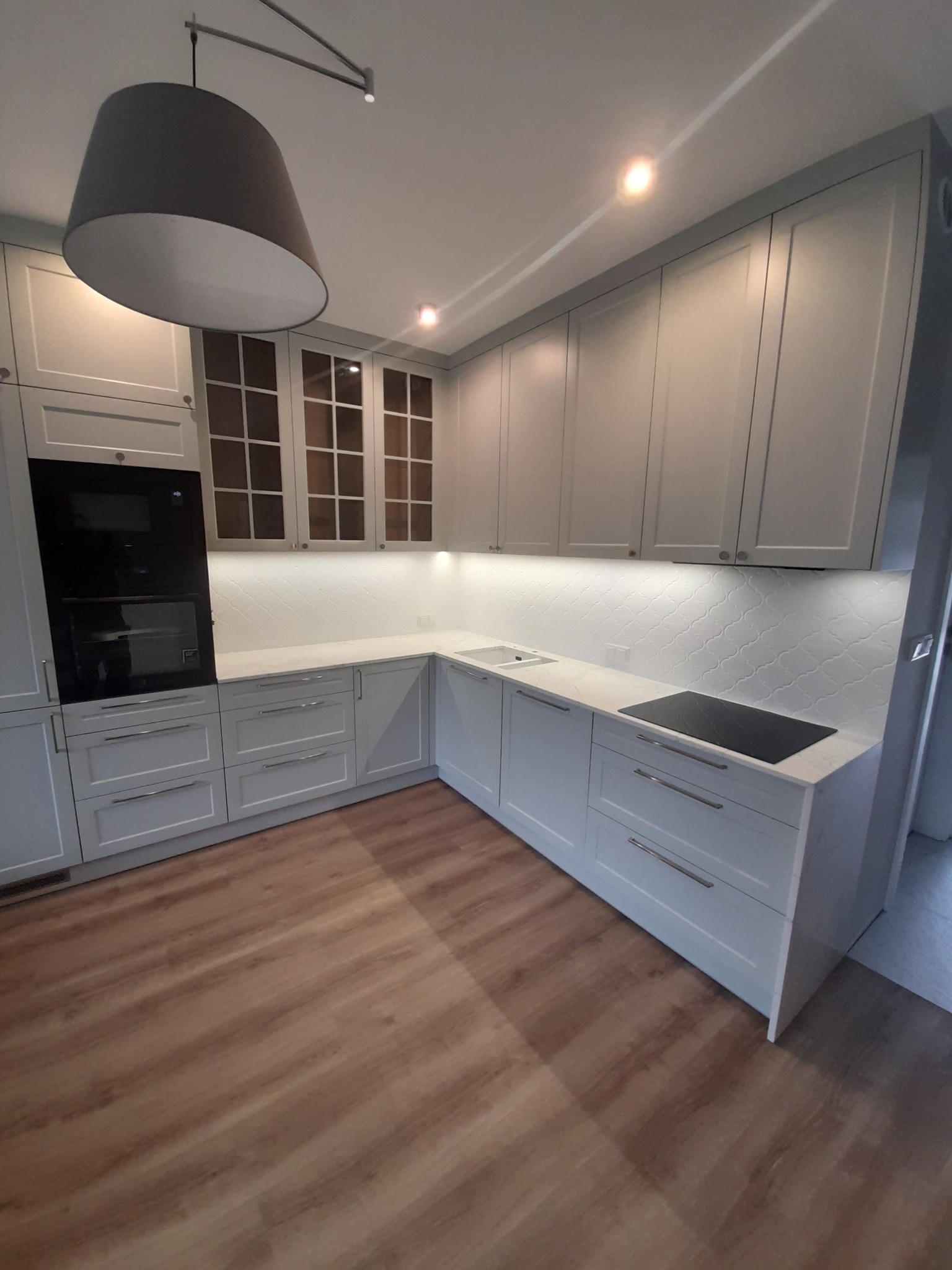 Jasna kuchnia z szarymi szafkami, białym blatem i drewnopodobną podłogą, oświetlona lampą wiszącą i punktowymi światłami podszafkowymi.