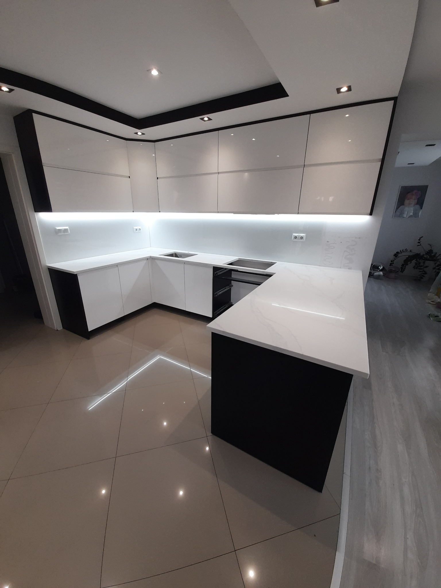 Nowoczesna kuchnia z białymi lakierowanymi szafkami i czarnymi akcentami, oświetlenie LED pod szafkami wiszącymi, jasny blat z delikatnym wzorem, błyszcząca podłoga.
