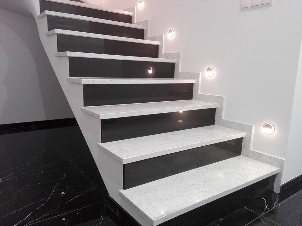 Nowoczesne schody z białego kamienia z czarnymi wstawkami, oświetlone punktowymi lampami LED na ścianie, wykończone czarną marmurową podłogą.