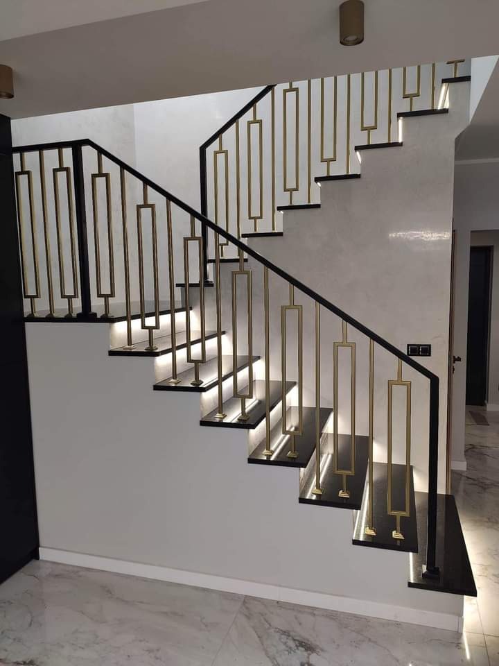 Nowoczesne schody z czarnymi stopniami, podświetleniem LED i złotymi balustradami o geometrycznym wzorze, widok z dołu na marmurowej podłodze.