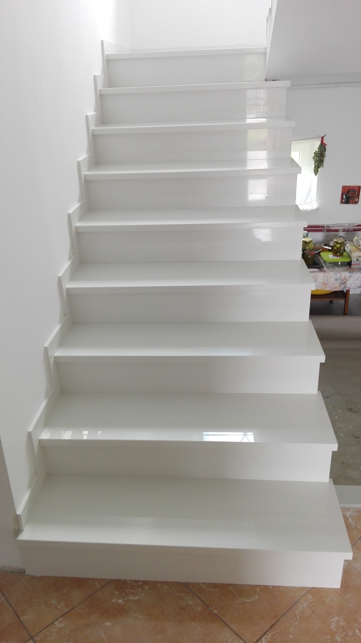 Białe, drewniane schody wewnętrzne z widocznymi stopniami i podstopnicami, wykończone na wysoki połysk, z brązową podłogą w tle.