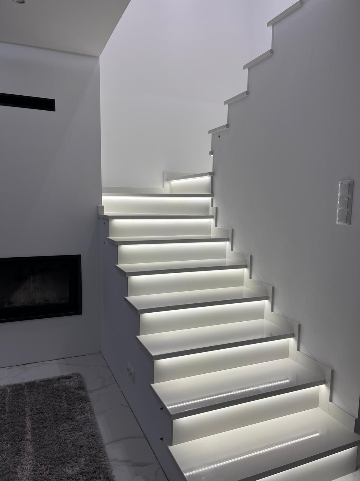 Białe schody z podświetleniem LED na każdym stopniu, obok kominek i szary dywan, minimalistyczny design.