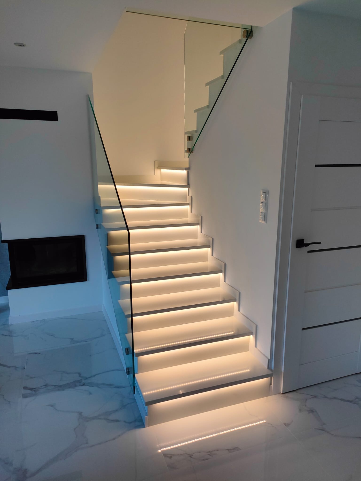 Nowoczesne schody wewnętrzne z podświetleniem LED stopni i szklaną balustradą, marmurowa posadzka, minimalistyczny design, obok białe drzwi z czarną klamką i dekoracyjny kominek.