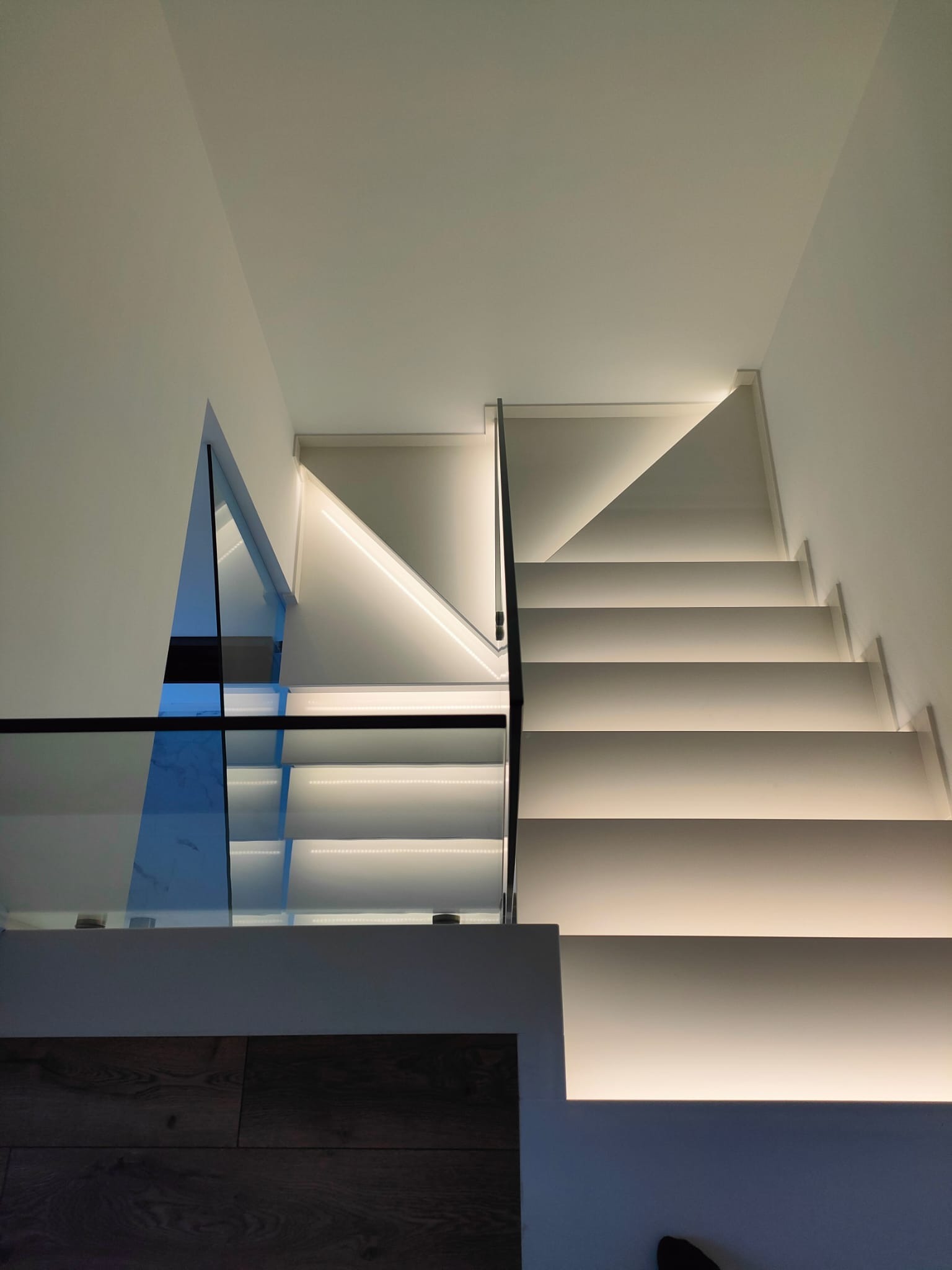 Nowoczesne, białe schody z podświetleniem LED i szklaną balustradą, widok od dołu, akcenty ciemnego drewna na podłodze.