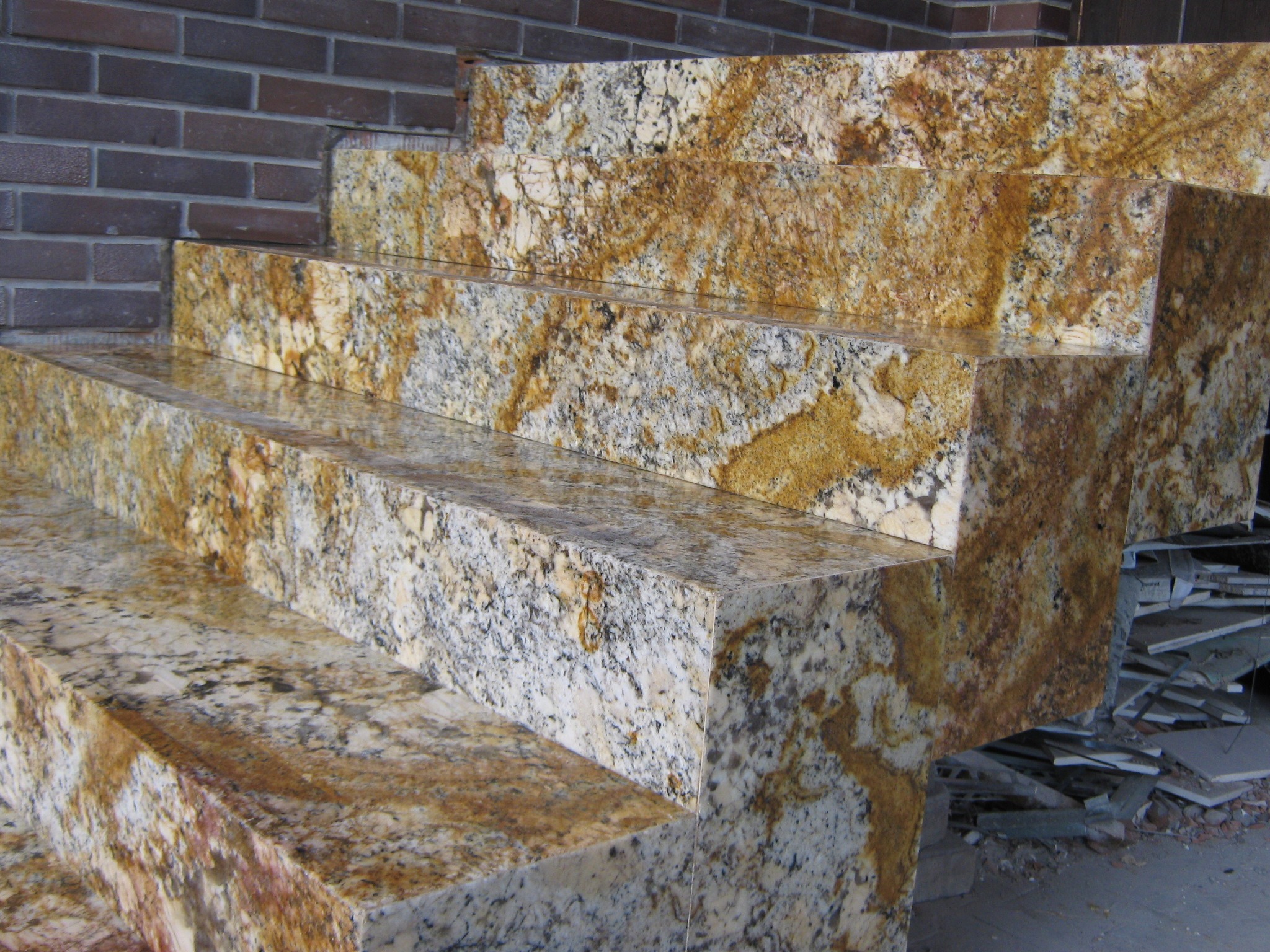 Wykończone zewnętrzne schody z polerowanego granitu w odcieniach złota i szarości, przylegające do ceglanej ściany, z widocznymi elementami gruzu po montażu.