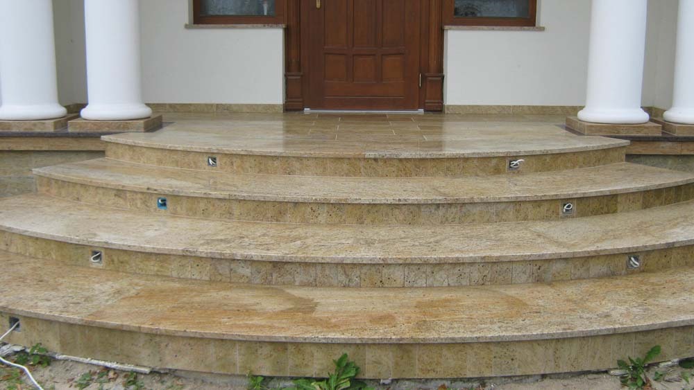 Wykończone, zaokrąglone schody wejściowe z jasnego kamienia z wbudowanym oświetleniem punktowym, prowadzące do drewnianych drzwi wejściowych i białych kolumn.