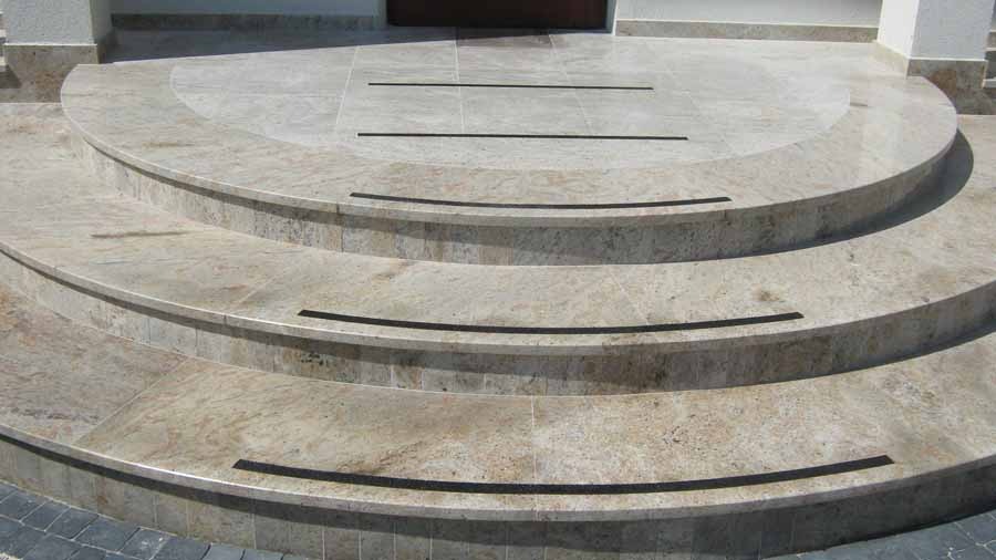 Półokrągłe schody zewnętrzne wyłożone beżowym kamieniem z ciemnymi paskami antypoślizgowymi, prowadzące do drzwi wejściowych.