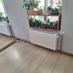 Biały grzejnik panelowy zamontowany na ścianie pod oknem z półkami pełnymi roślin doniczkowych, widoczne miedziane rury instalacji grzewczej i fragment podłogi z rozwiniętym kartonem ochronnym...