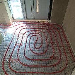 PARUS Instalacje Hydrauliczne Paweł Rusowicz - Czerwone rury ogrzewania podłogowego marki Roth, ułożone spiralnie na srebrnej macie z siatką, widoczne okno w tle. Gotowa instalacja przed wylaniem posadzki.