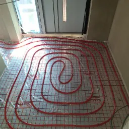 Czerwone rury ogrzewania podłogowego marki Roth, ułożone spiralnie na srebrnej macie z siatką, widoczne okno w tle. Gotowa instalacja przed wylaniem posadzki.