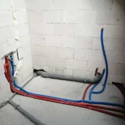 Surowe wnętrze łazienki z widoczną instalacją wodno-kanalizacyjną: rury PCV szare i rury PEX czerwone i niebieskie, mocowane do ściany z betonu komórkowego, częściowo wypełnionej pianką montażową.