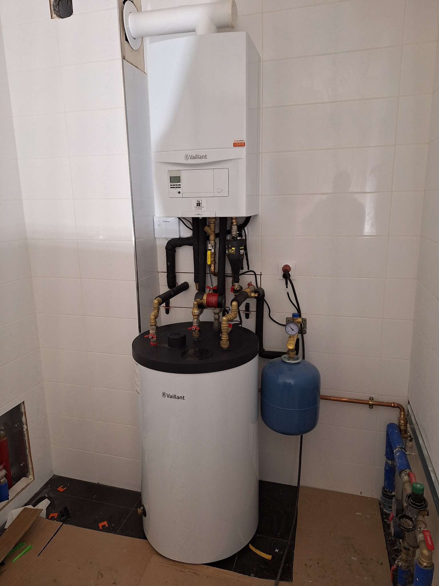 Nowoczesny, biały piec gazowy Vaillant z zasobnikiem wody i niebieskim naczyniem przeponowym, zainstalowany na tle białych płytek ceramicznych, widoczne elementy instalacji hydraulicznej...