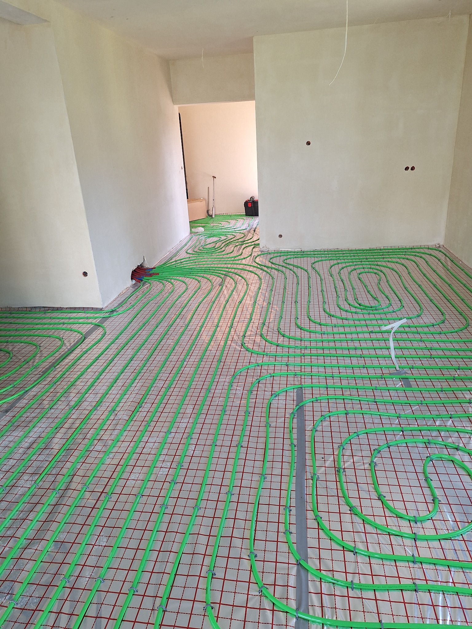 Świeżo ułożone, zielone rurki ogrzewania podłogowego na czerwonej siatce w surowym wnętrzu, z widocznym kolektorem przy ścianie i narzędziami w tle.