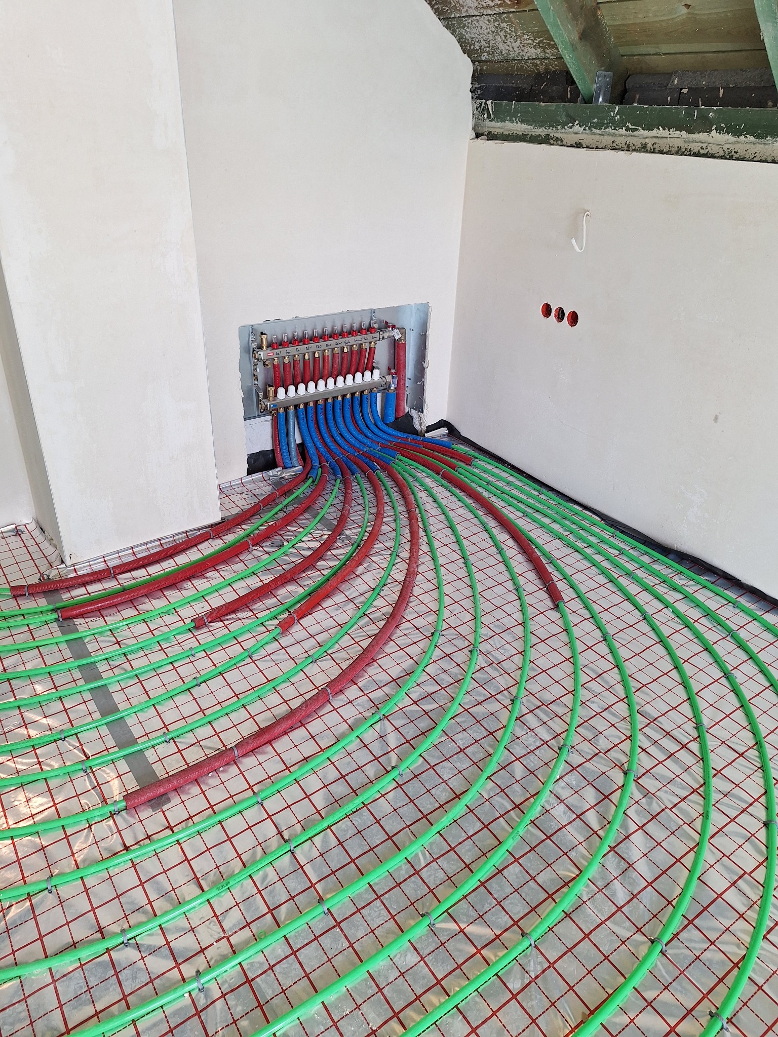 Rozdzielacz ogrzewania podłogowego z podłączonymi rurami w kolorach czerwonym, niebieskim i zielonym, ułożonymi na folii z siatką w nowym budynku w trakcie instalacji.