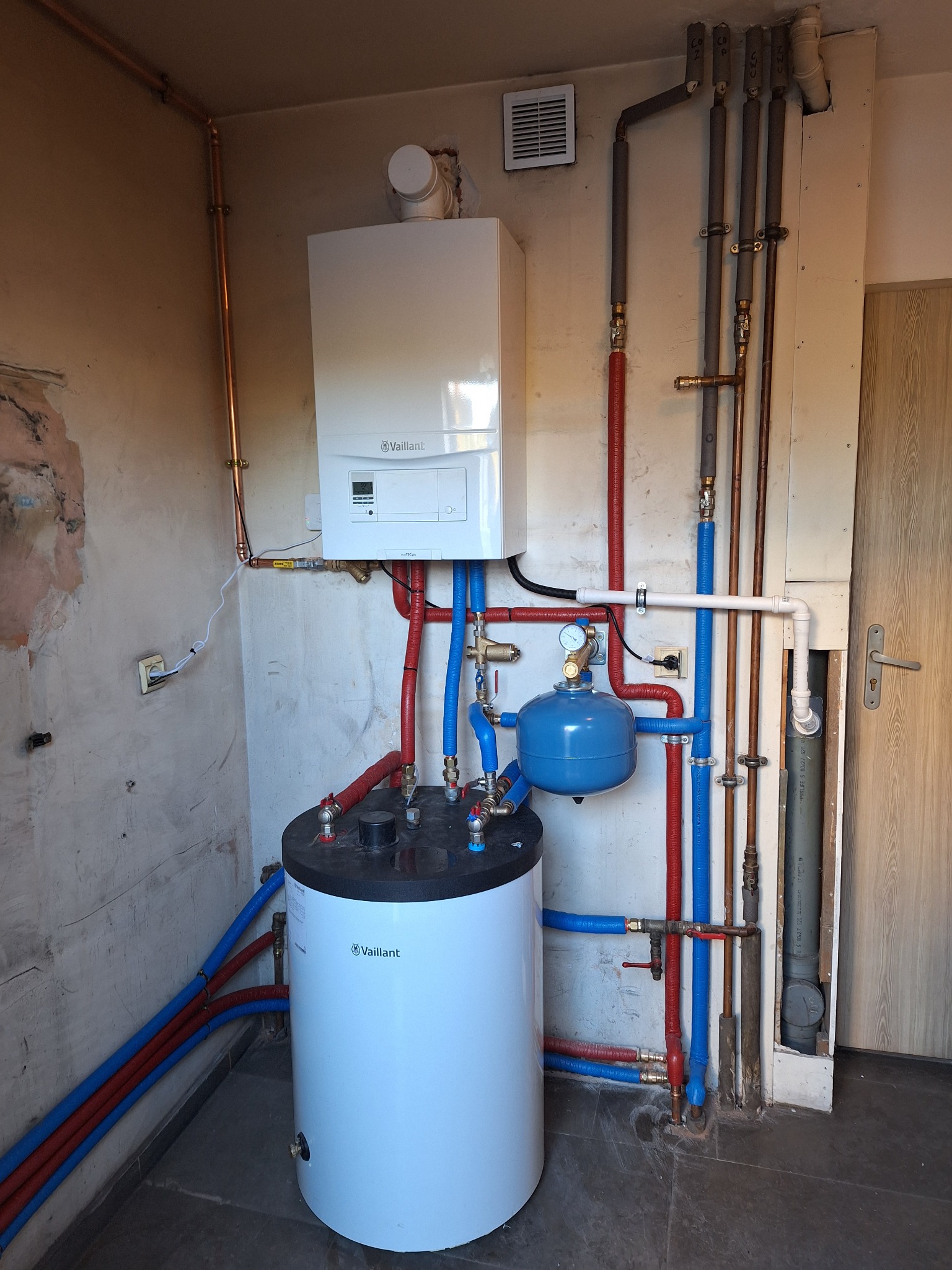 Kompletna instalacja grzewcza z piecem gazowym i zasobnikiem wody, widoczne rury miedziane i plastikowe, elementy armatury, zamontowany piec gazowy marki Vaillant, nieotynkowana ściana w tle.