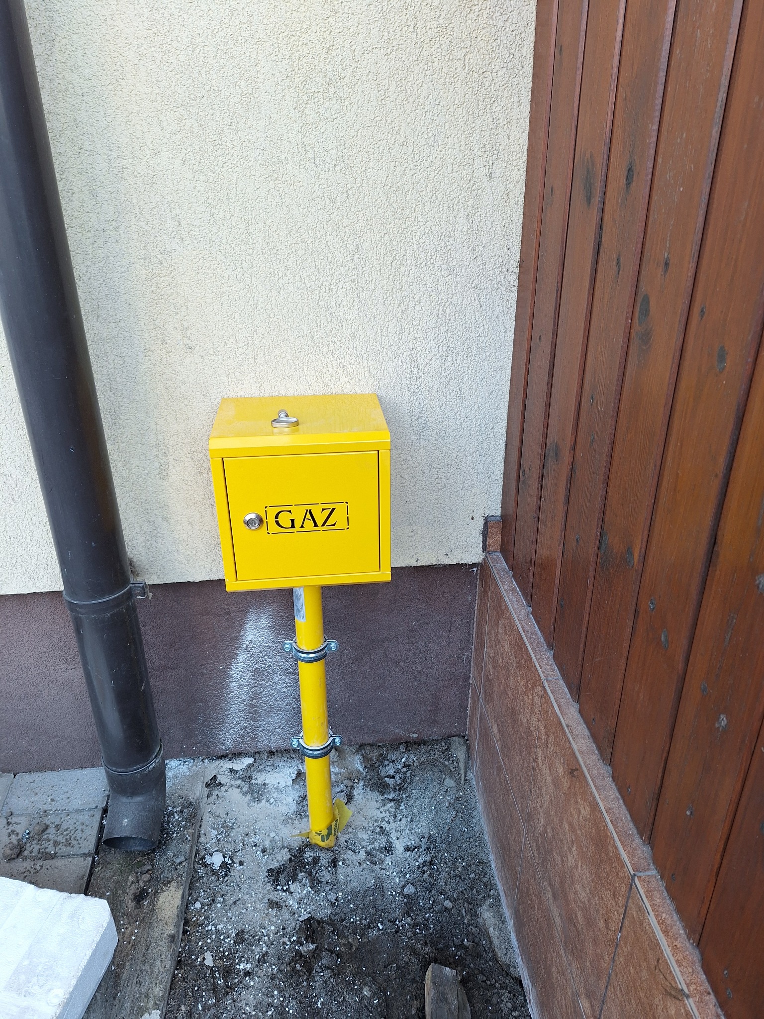 Żółta skrzynka gazowa z napisem GAZ, zamontowana na rurze, widoczna obok rury spustowej i drewnianego ogrodzenia.