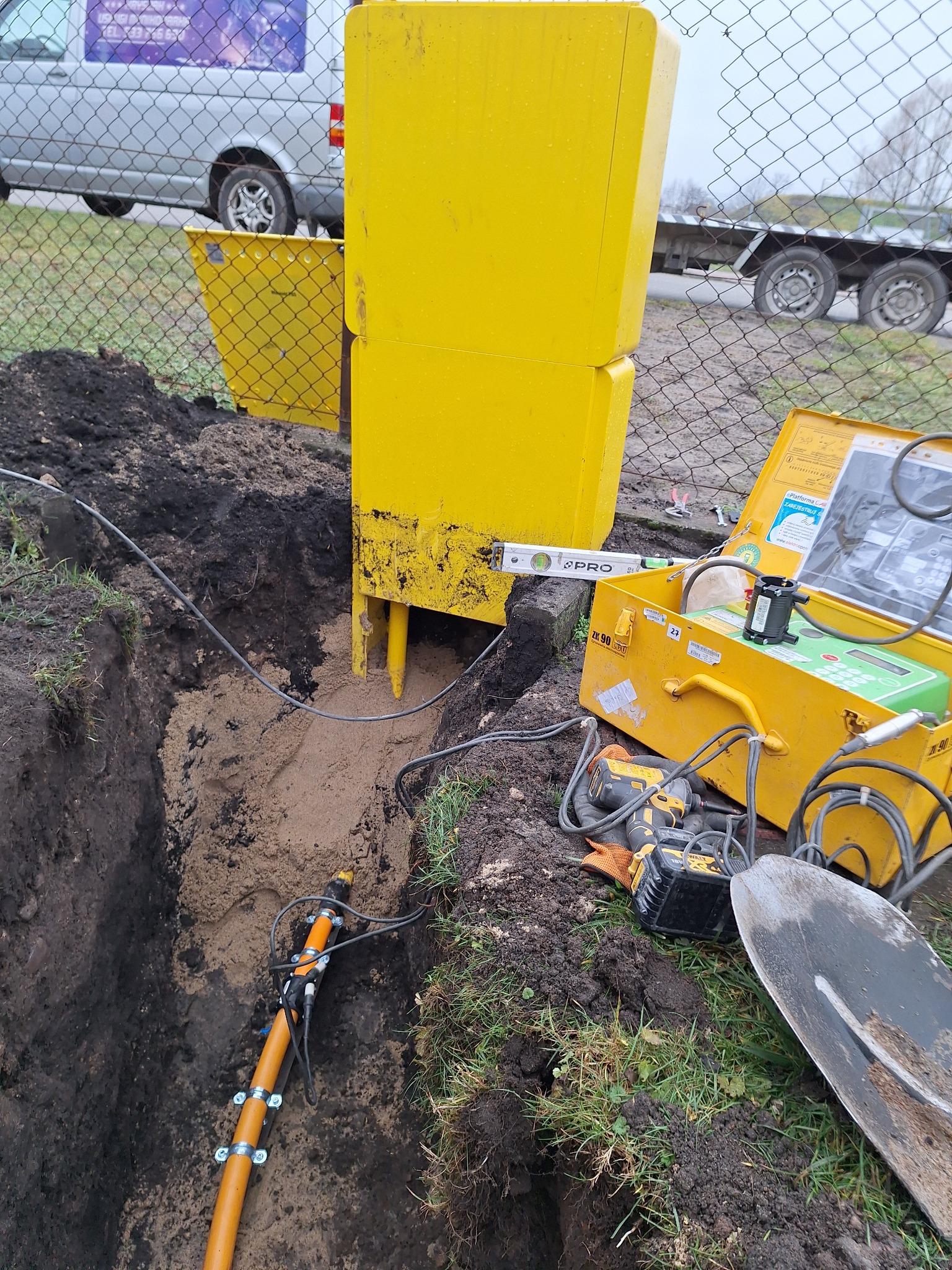Żółta skrzynka zabezpieczająca nad wykopem z rurą gazową, poziomica, otwarty żółty kufer z urządzeniami pomiarowymi, wiertarka akumulatorowa i łopata na trawie, w tle ogrodzenie i samochód.