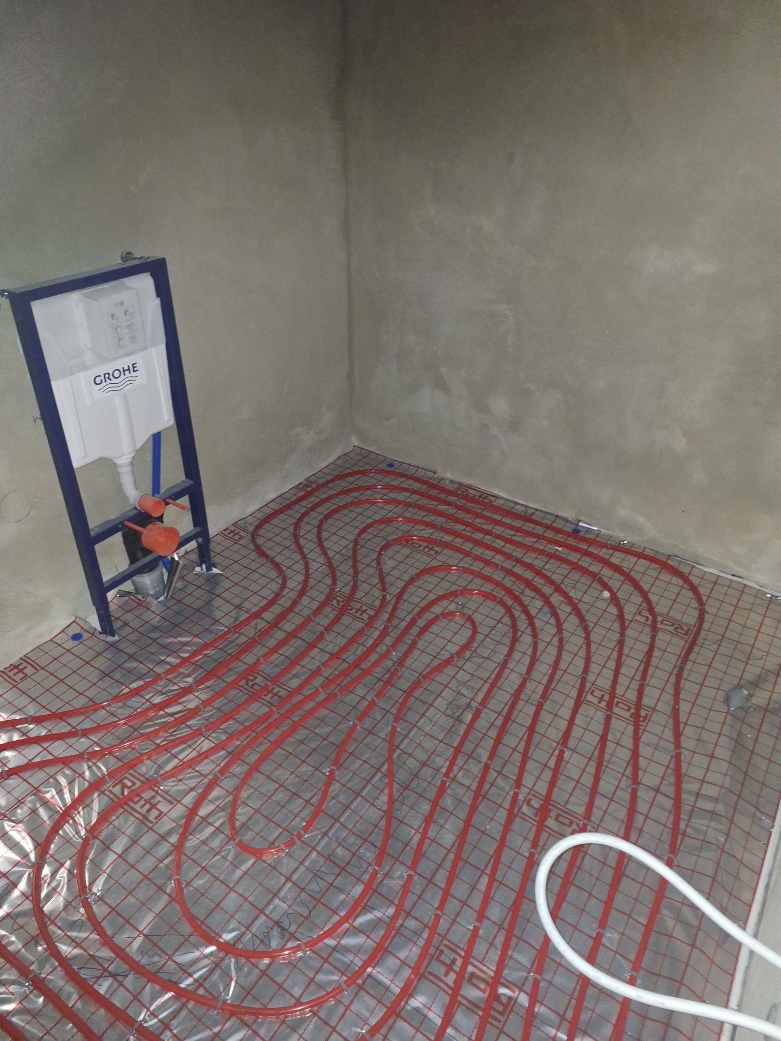 Czerwone rury ogrzewania podłogowego ułożone na srebrnej macie w surowym wnętrzu łazienki, z widocznym stelażem podtynkowym Grohe.