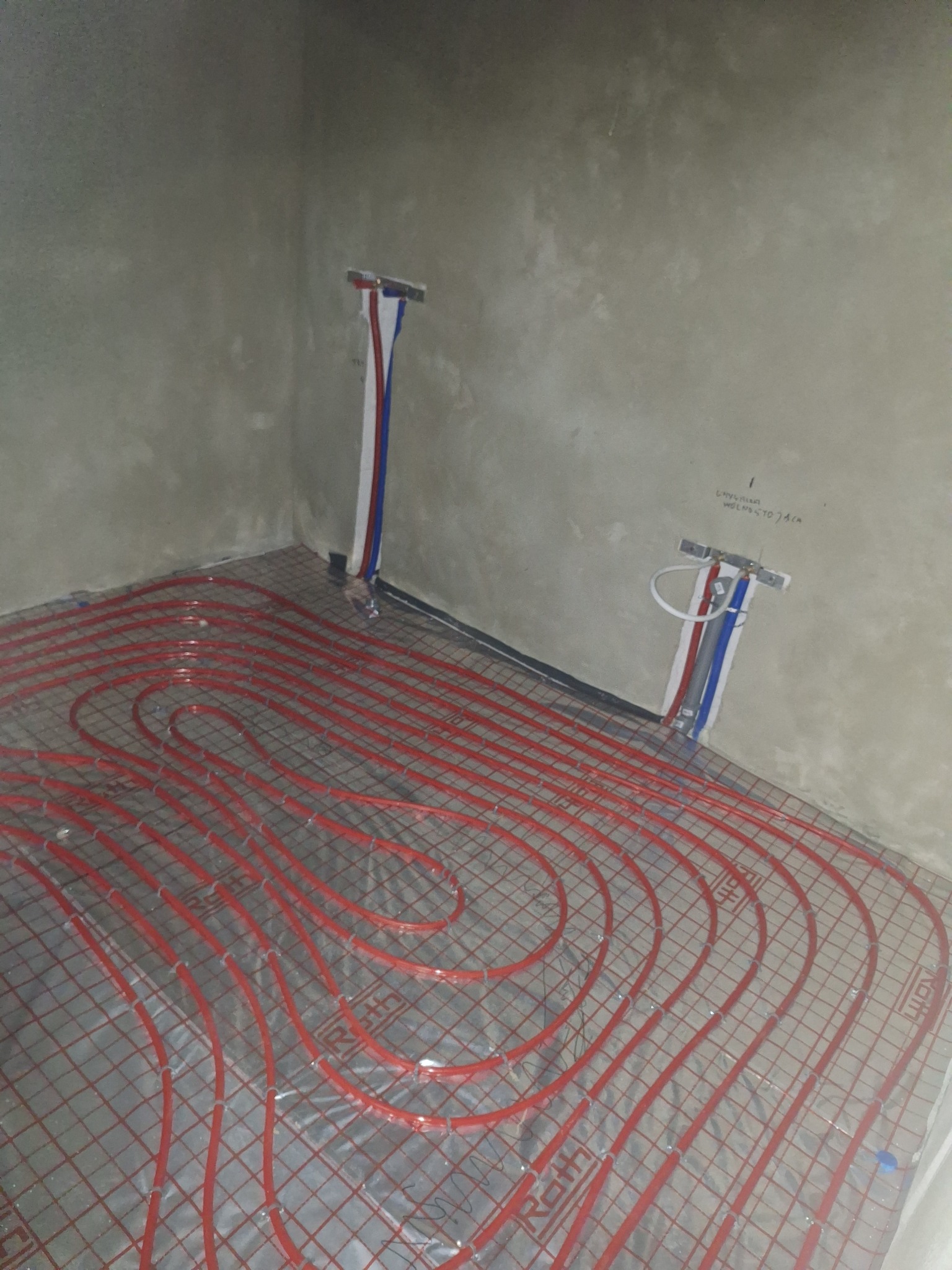 Czerwone rury ogrzewania podłogowego ułożone na srebrnej folii, widoczne przyłącza do instalacji na ścianie w surowym pomieszczeniu.