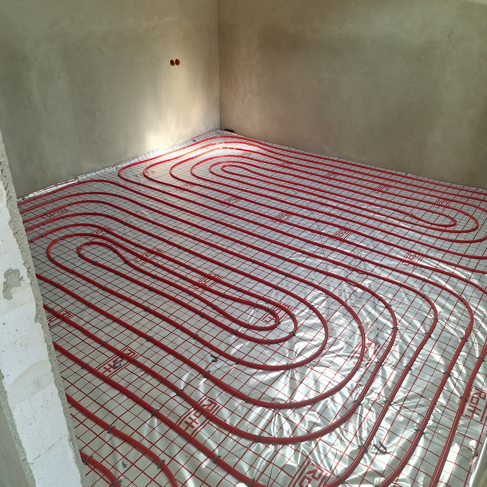 Czerwone rury ogrzewania podłogowego ułożone spiralnie na folii izolacyjnej w surowym pomieszczeniu. Widok z góry, etap instalacji przed wylewką.