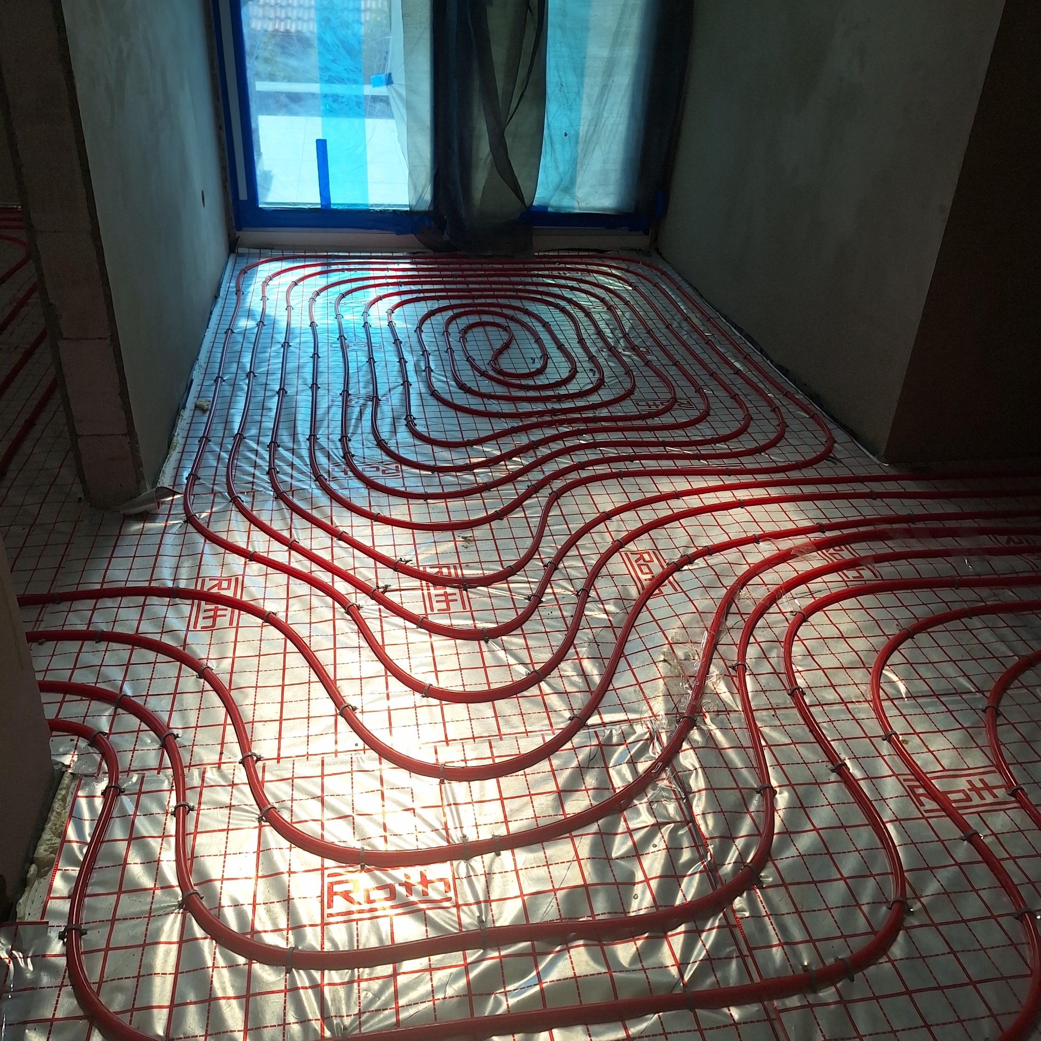 Czerwone rury ogrzewania podłogowego ułożone na srebrnej folii z siatką, widok z góry, okno w tle. System ogrzewania podłogowego marki Roth w surowym wnętrzu.