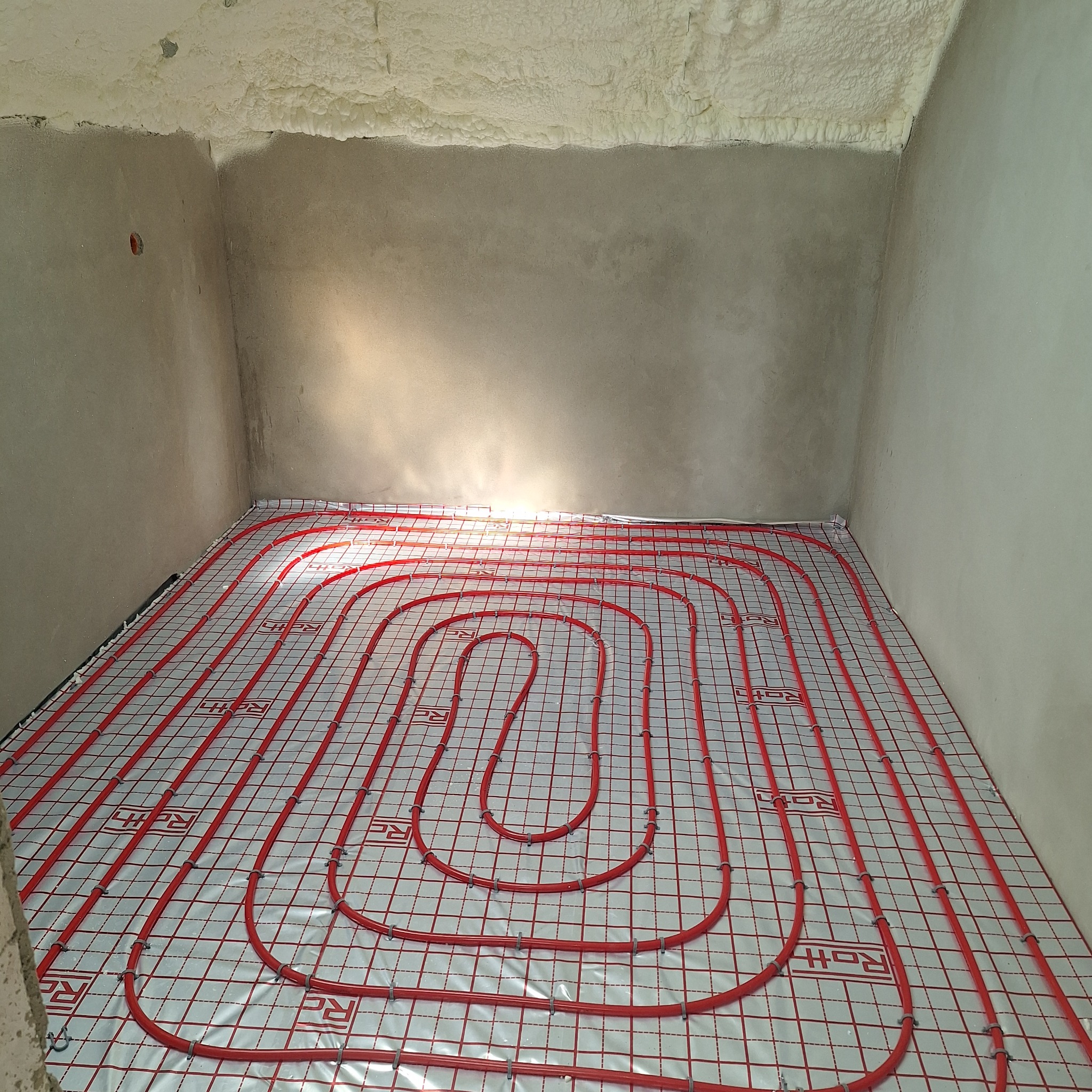Czerwone rury ogrzewania podłogowego ułożone na folii z rastrem w surowym wnętrzu, ściany w szarym tynku, sufit z pianki montażowej.