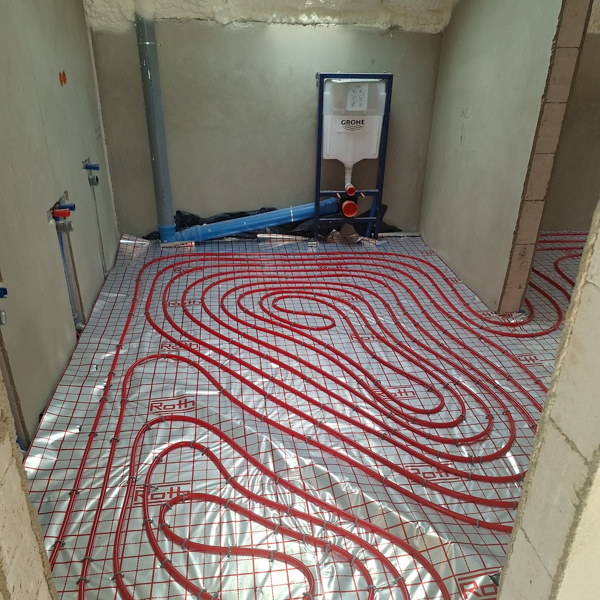 Instalacja ogrzewania podłogowego Roth w surowym wnętrzu. Czerwone rury ułożone na srebrnej folii, widoczny zbiornik Grohe i fragment instalacji wodnej na ścianie.