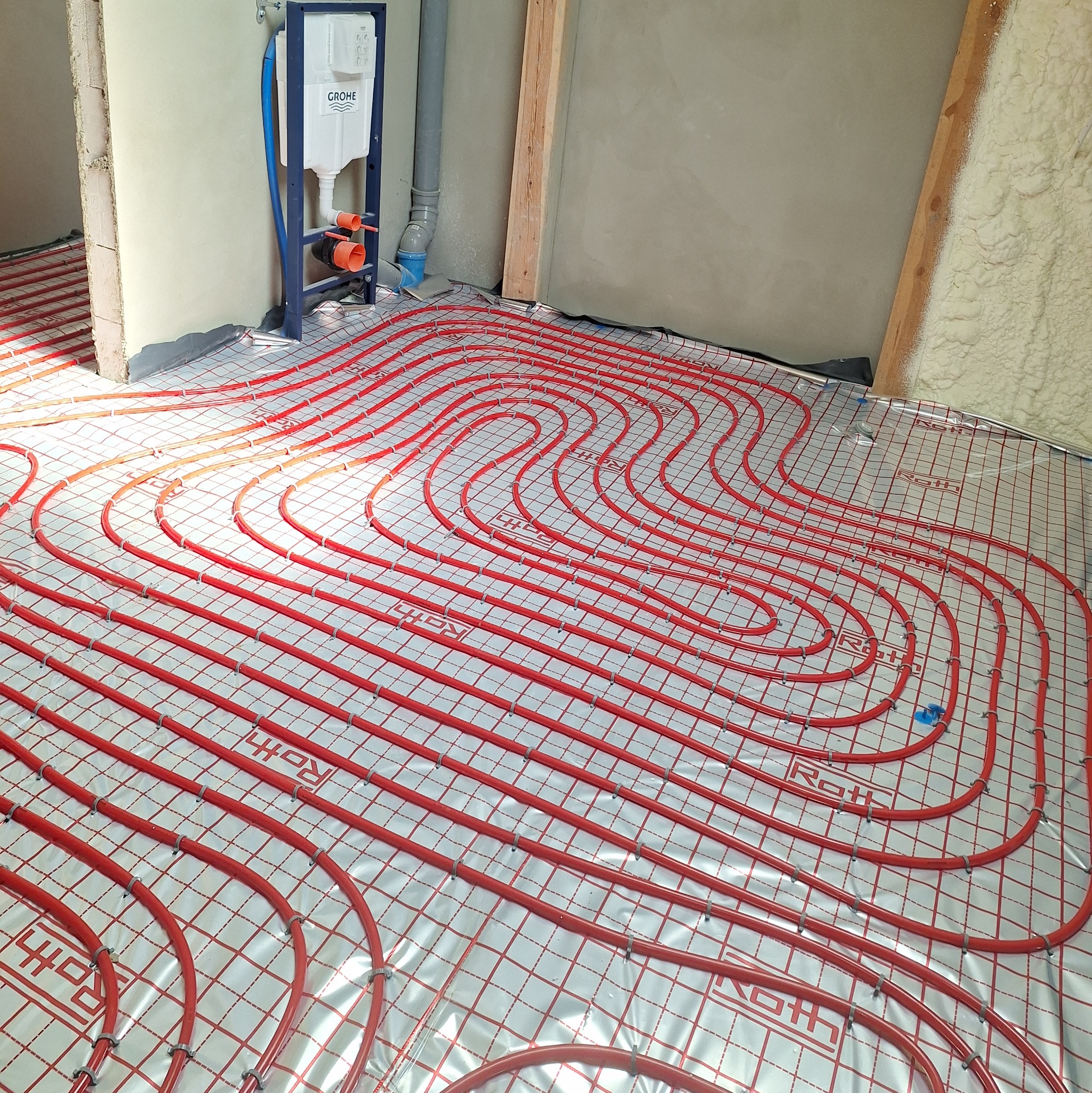 Czerwone rury ogrzewania podłogowego Roth ułożone na folii z nadrukiem kratki, widoczny stelaż podtynkowy WC Grohe, fragment ściany z pianką izolacyjną.