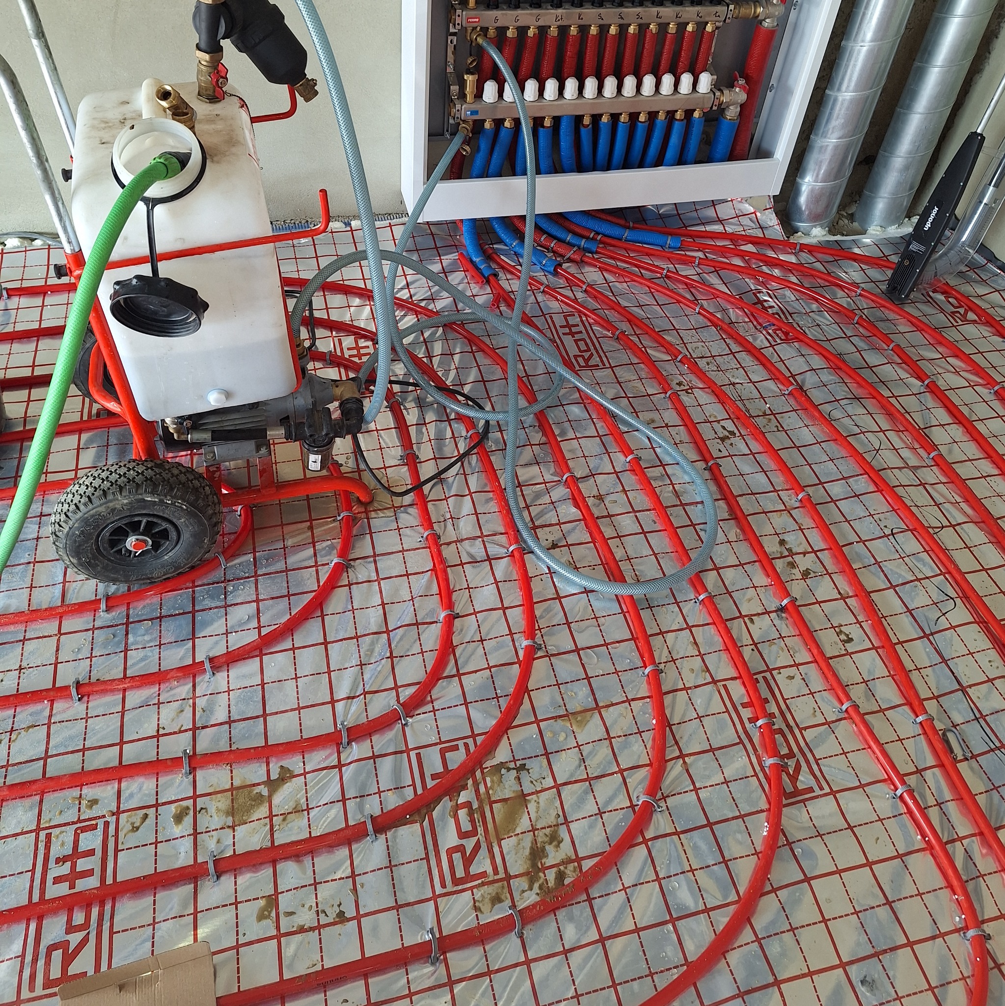 Czerwone rury ogrzewania podłogowego na folii, z podłączonym rozdzielaczem i maszyną do napełniania. Widoczne rury wentylacyjne i narzędzie pomiarowe Upornor.