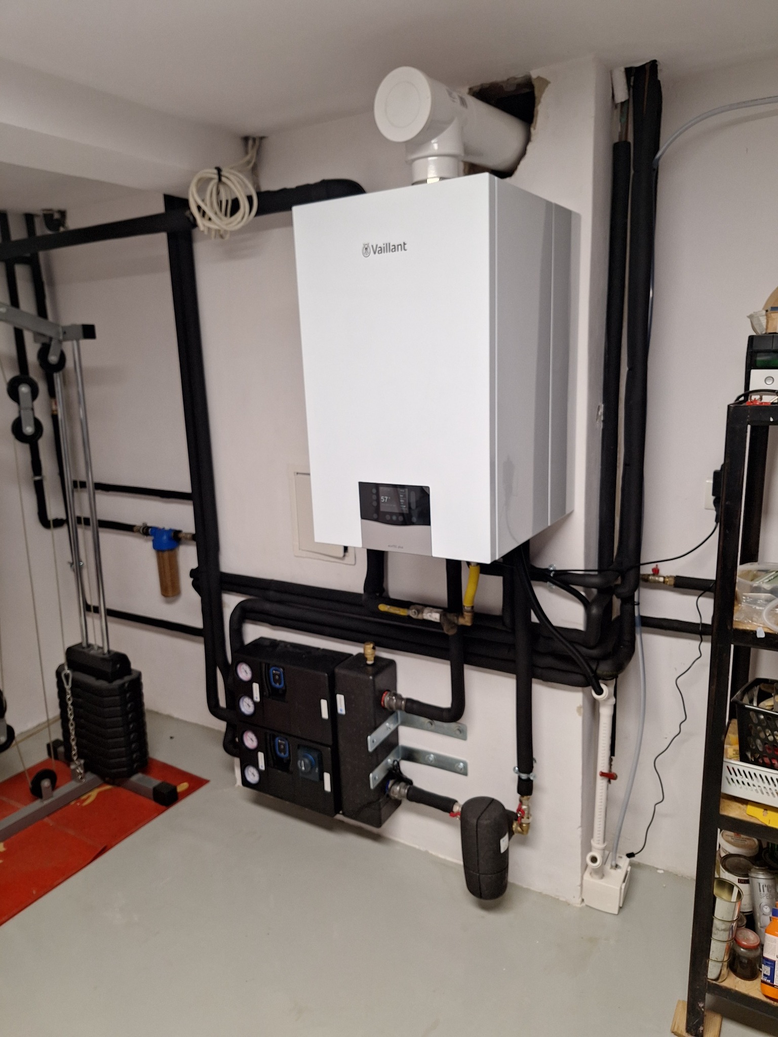 Biały piec gazowy Vaillant z podłączeniami na tle ściany, widoczne elementy instalacji i rury. W tle fragment siłowni domowej.
