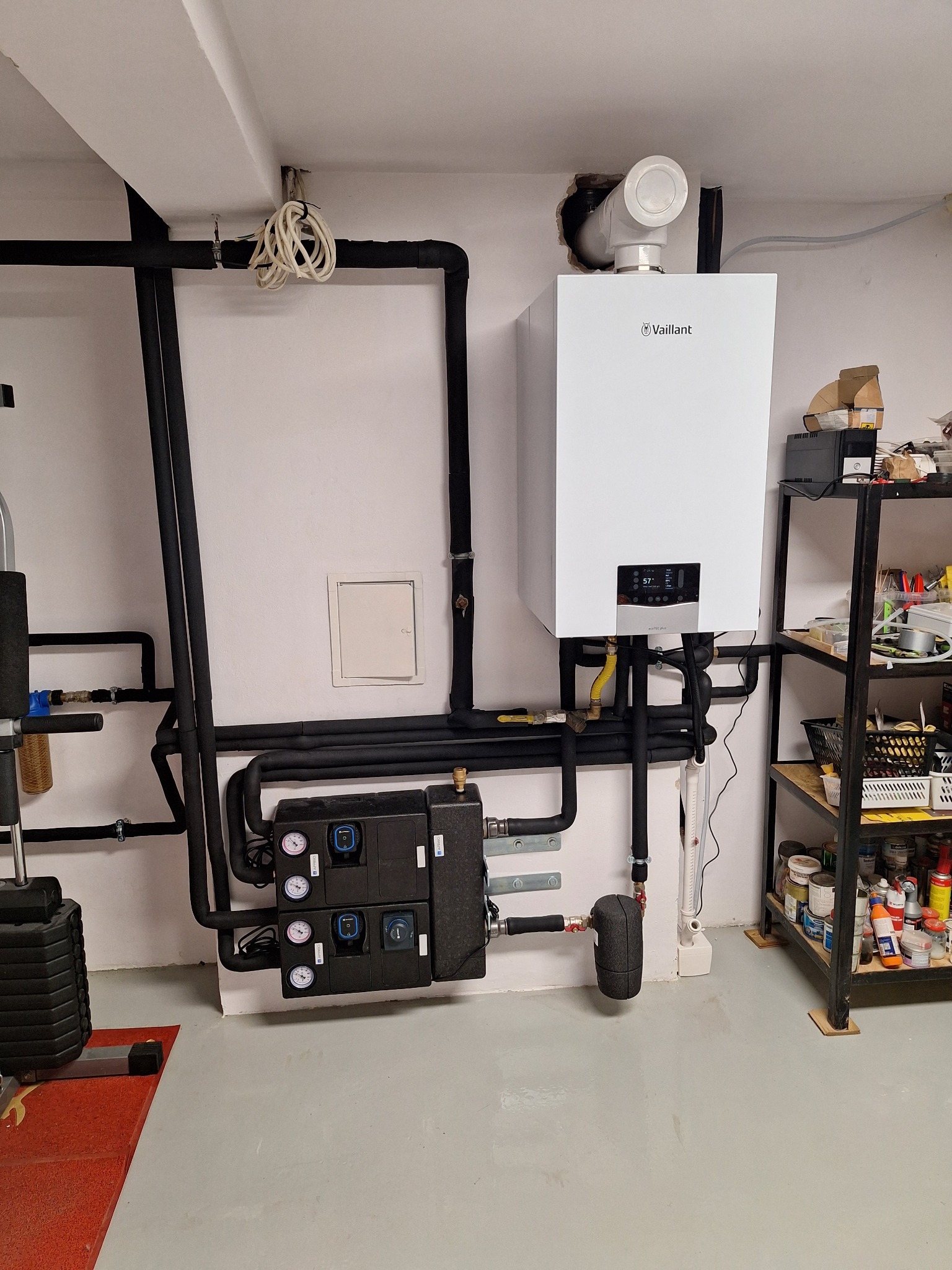 Nowoczesny, biały piec gazowy Vaillant zamontowany na ścianie z czarnymi rurami instalacji grzewczej. Obok regał z narzędziami i akcesoriami. Widoczny sprzęt do ćwiczeń.