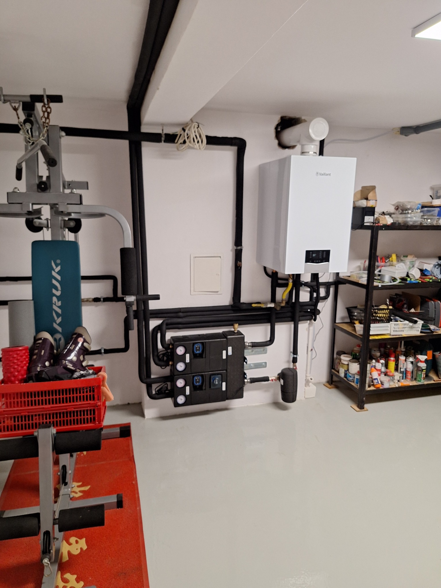 Biały piec gazowy Vaillant z czarną instalacją w garażu, obok regał z narzędziami i sprzęt do ćwiczeń. Rury prowadzone po ścianie i suficie.