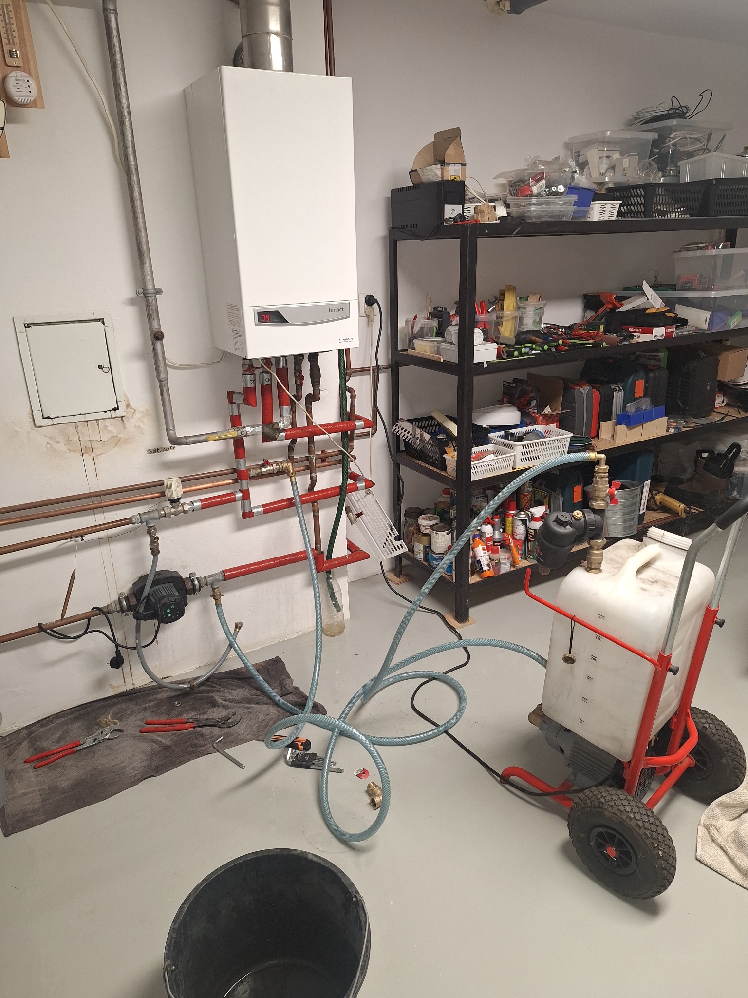 Biały piec gazowy Termet na ścianie z podłączonymi rurami miedzianymi i czerwonymi, obok regał z narzędziami, w tle biała ściana, widoczne narzędzia hydrauliczne.