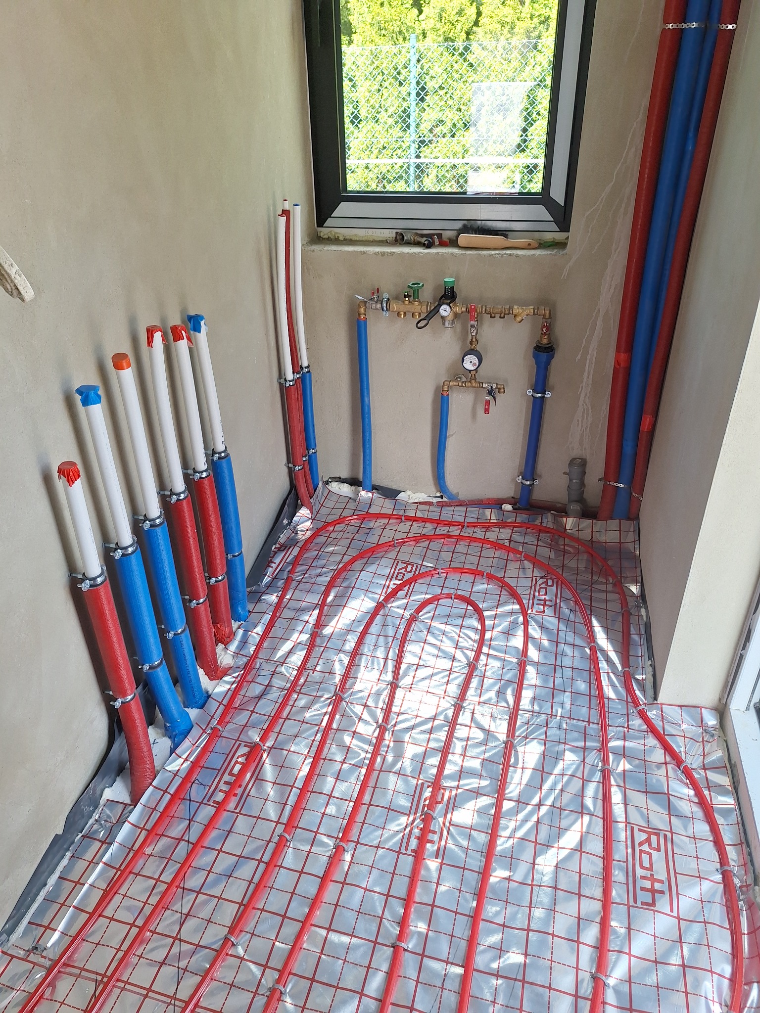 Instalacja ogrzewania podłogowego z czerwonymi rurkami na srebrnej folii, widok z góry, rury pionowe z niebiesko-czerwonymi osłonami, okno w tle.