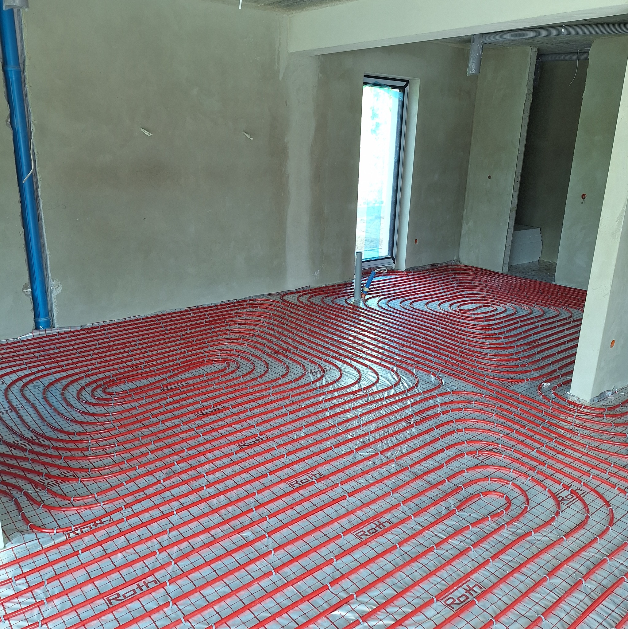 Czerwone rury ogrzewania podłogowego marki Roth ułożone spiralnie na folii, w surowym wnętrzu z oknem i nieotynkowanymi ścianami, gotowe do zalania wylewką.