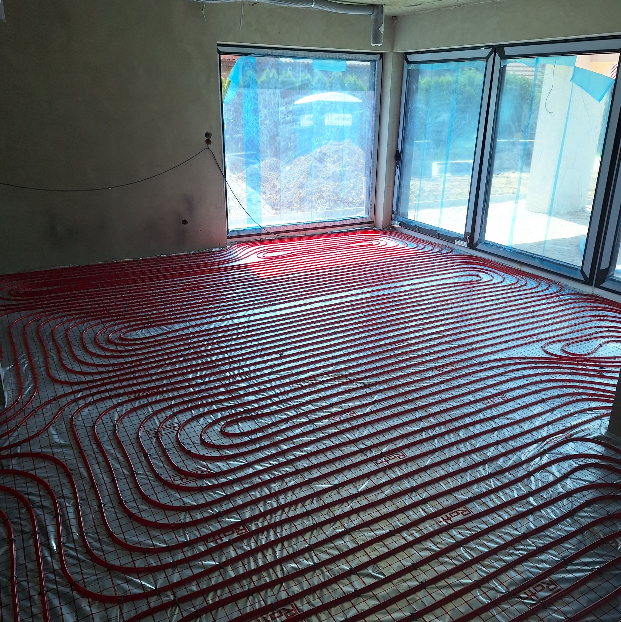 Czerwone rury ogrzewania podłogowego ułożone na srebrnej folii izolacyjnej w jasnym pomieszczeniu z dużymi oknami, przygotowane do zalania wylewką.