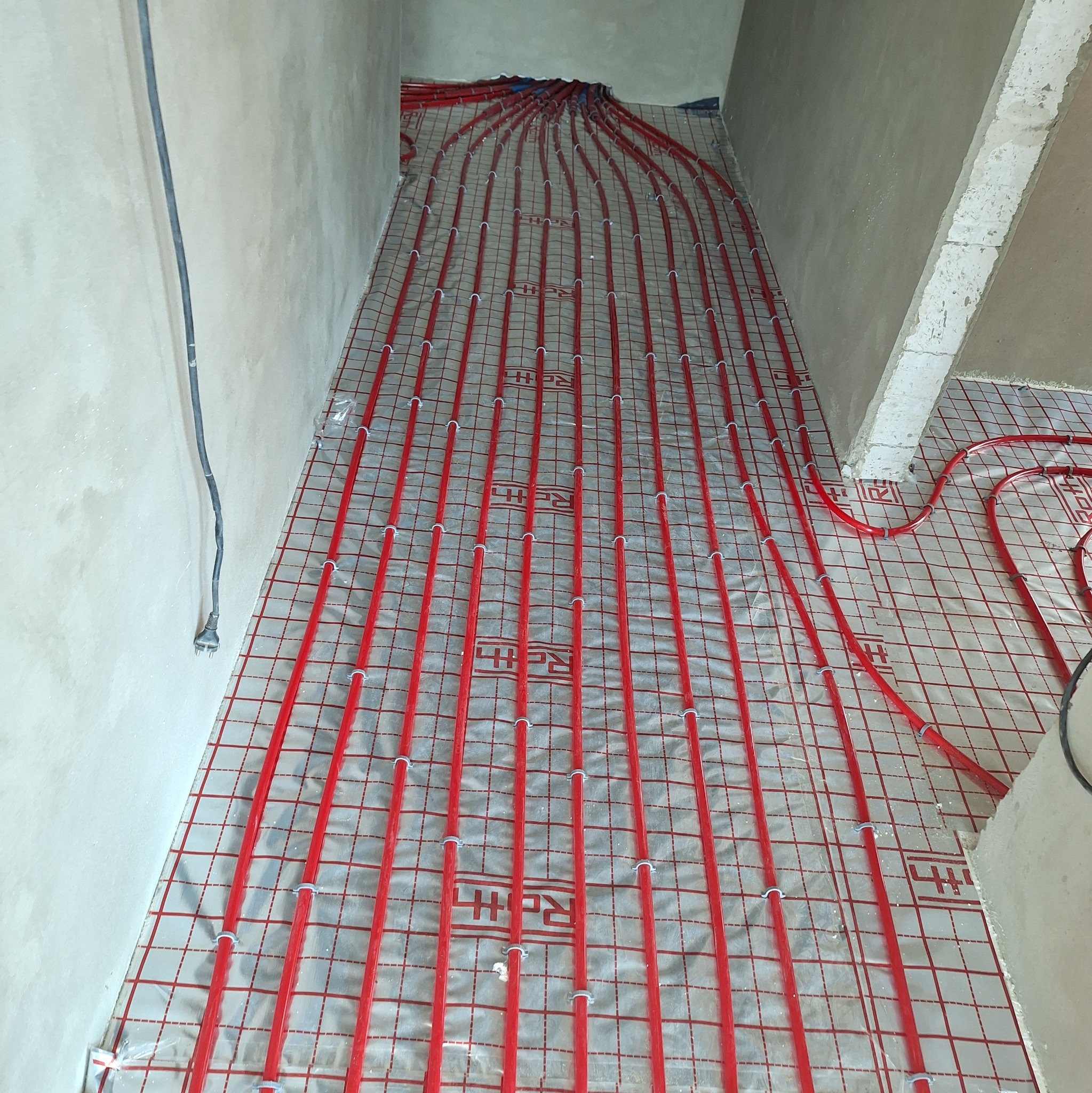 Czerwone rury ogrzewania podłogowego ułożone na izolacji, przymocowane białymi uchwytami, widoczne surowe ściany w trakcie remontu.