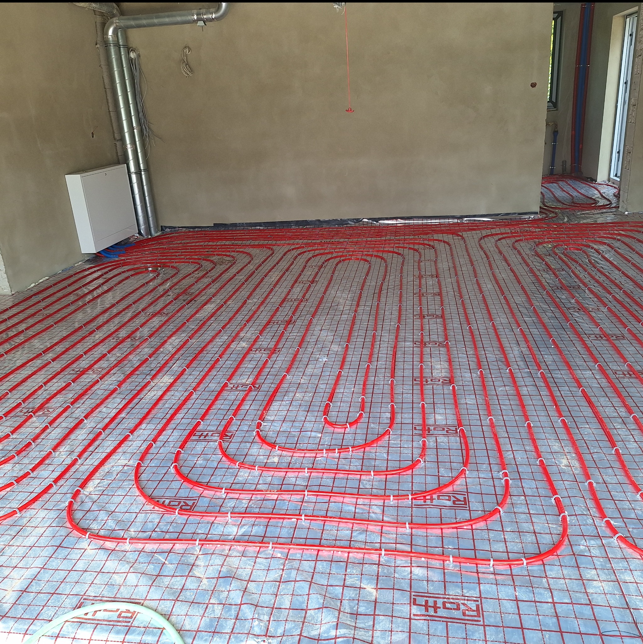 Czerwone rury ogrzewania podłogowego ułożone na srebrnej folii z siatką w surowym wnętrzu. Widoczne instalacje na ścianach i suficie.