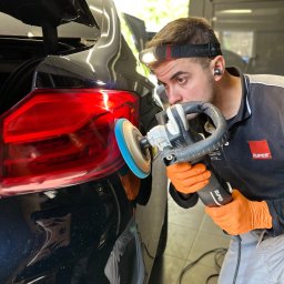 DYTKARS AutoDetailing - Detailer w pomarańczowych rękawicach poleruje tylną lampę czarnego samochodu przy użyciu polerki Rupes, oświetlając sobie pracę lampą czołową.