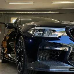 Błyszczący, czarny samochód sportowy z karbonowymi elementami, sfotografowany pod niskim kątem w dobrze oświetlonym garażu z szarą podłogą i ścianami.