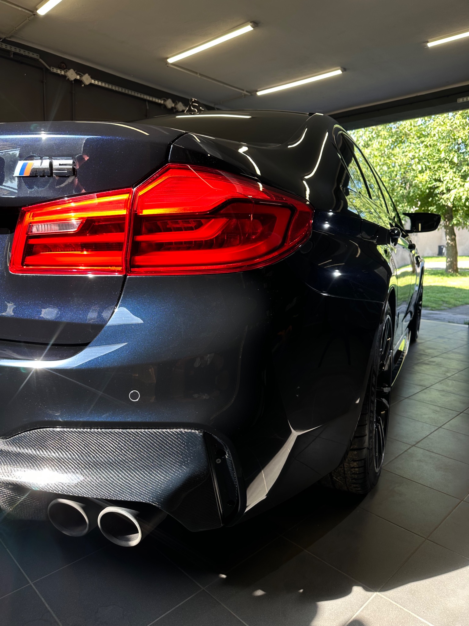 Błyszczący, granatowy samochód BMW M5 z widocznymi chromowanymi końcówkami wydechu i karbonowym dyfuzorem, zaparkowany w jasnym garażu z widokiem na zieleń na zewnątrz.