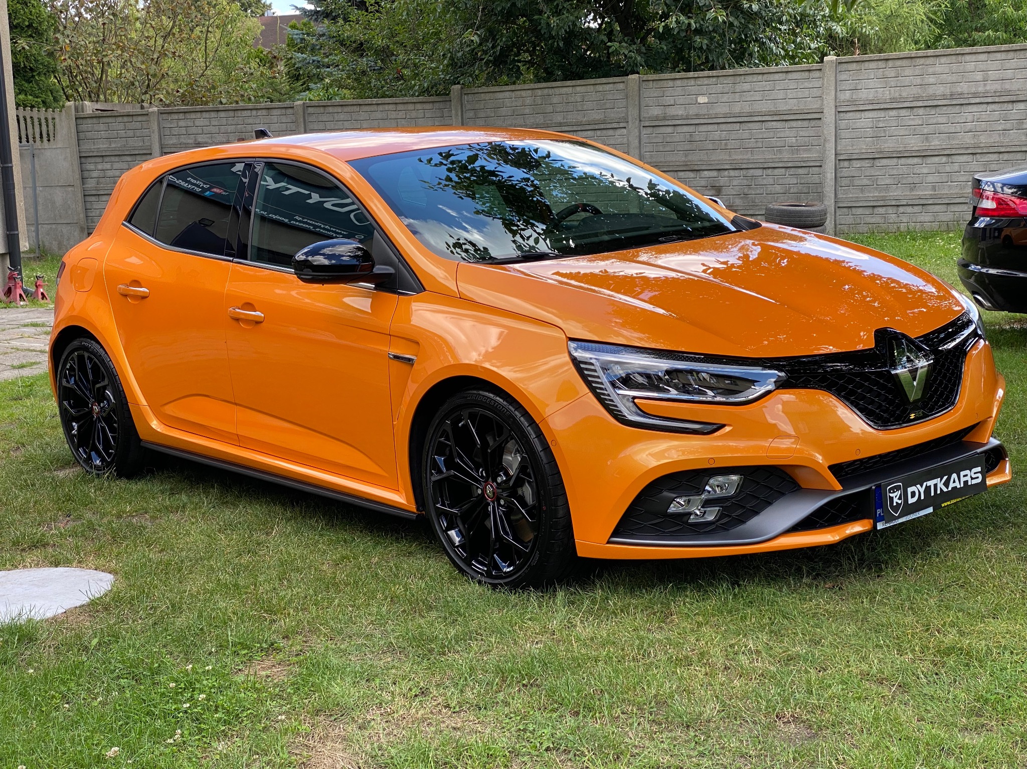 Pomarańczowy samochód Renault Megane RS z czarnymi felgami na trawniku przed betonowym ogrodzeniem.