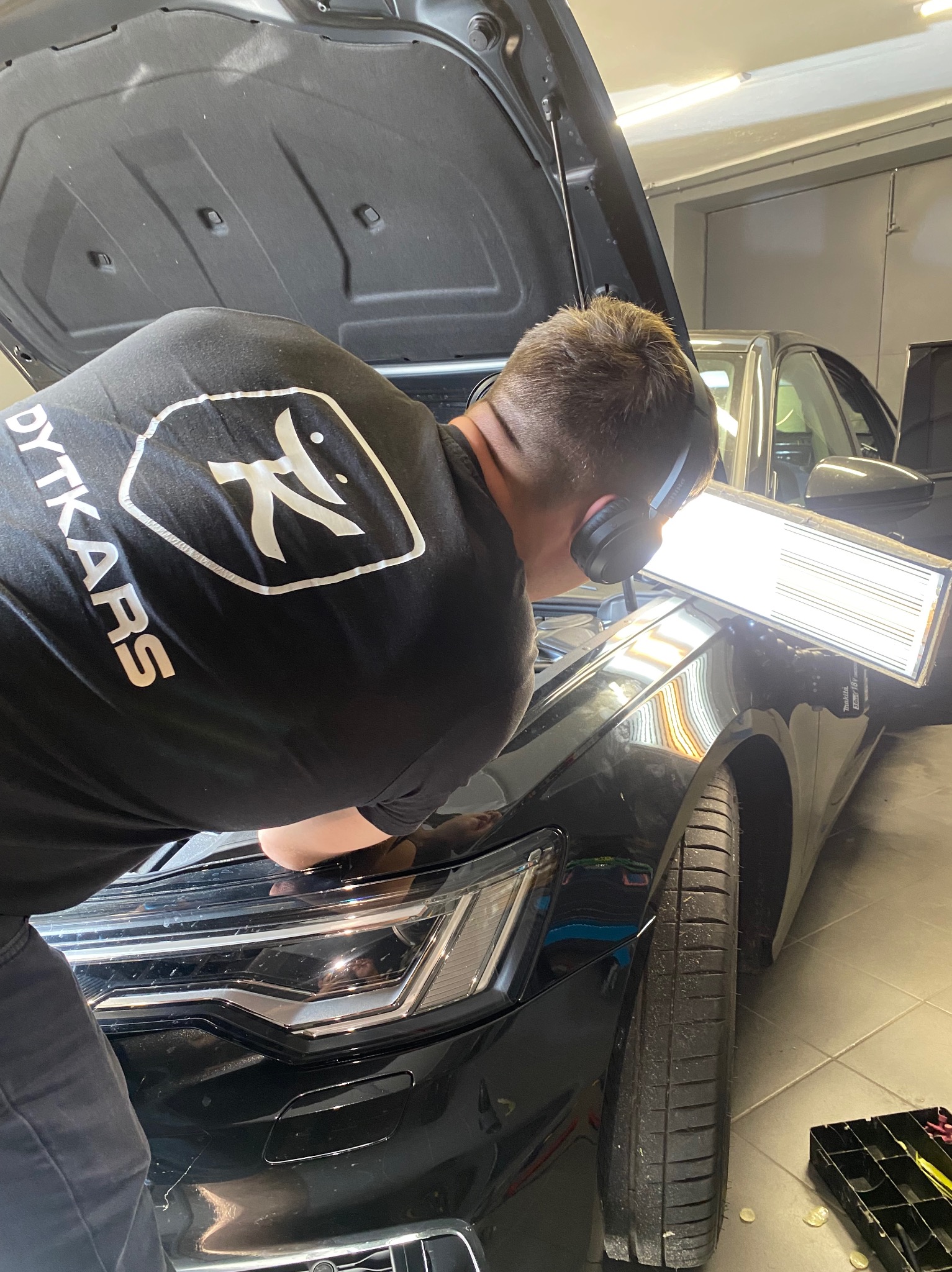 Mężczyzna w słuchawkach, ubrany w czarną koszulkę z logo firmy 'DYTKARS', pracuje nad karoserią czarnego samochodu z otwartą maską, używając specjalistycznej lampy inspekcyjnej do wykrywania...